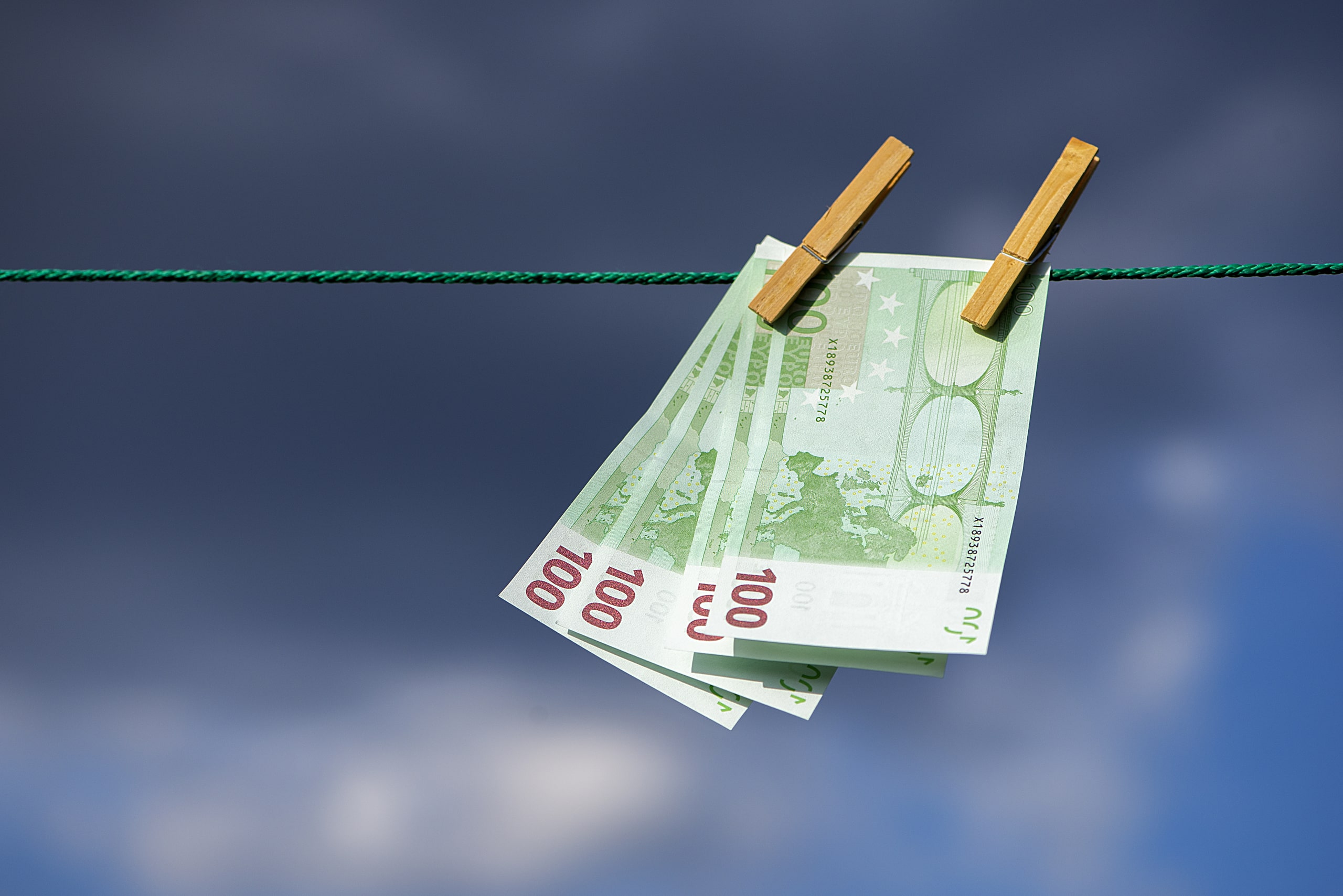 Prevención De Blanqueo De Capitales En La Auditoría Externa Billetes de 100 euros colgados en una cuerda con pinzas de madera, simbolizando el concepto de blanqueo de dinero en un cielo nublado.