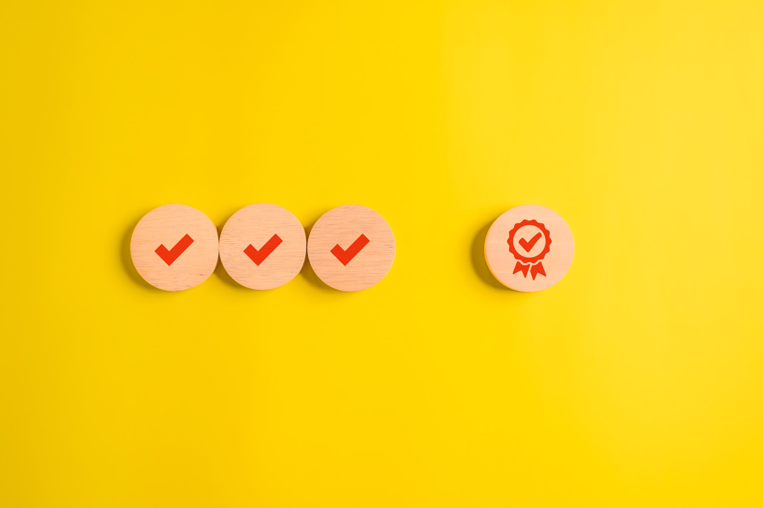 Elementos Clave En Los Procesos De Auditoría Forense Cuatro fichas de madera con marcas de verificación rojas y una ficha adicional con un icono de certificación, representando la validación y calidad en los procesos de auditoría forense sobre un fondo amarillo.