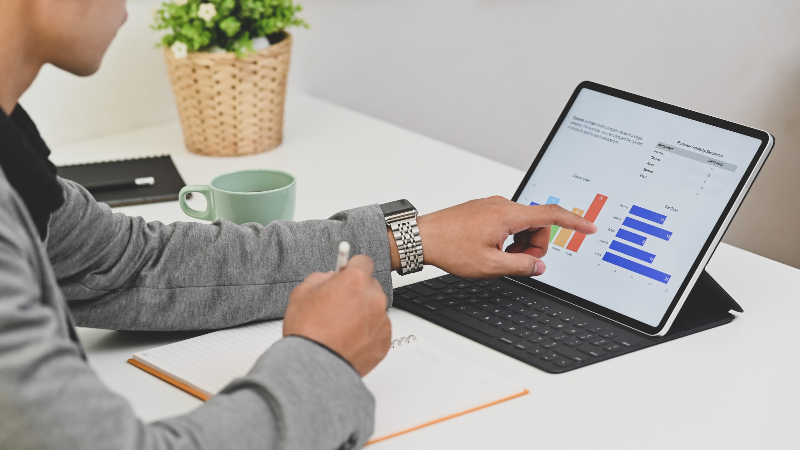 Análisis De Transacciones Revisión De Datos Financieros En Tiempo Real Persona realizando un análisis de transacciones en una tablet, observando gráficos de barras y datos financieros, mientras toma notas en una libreta en un entorno de oficina moderno.