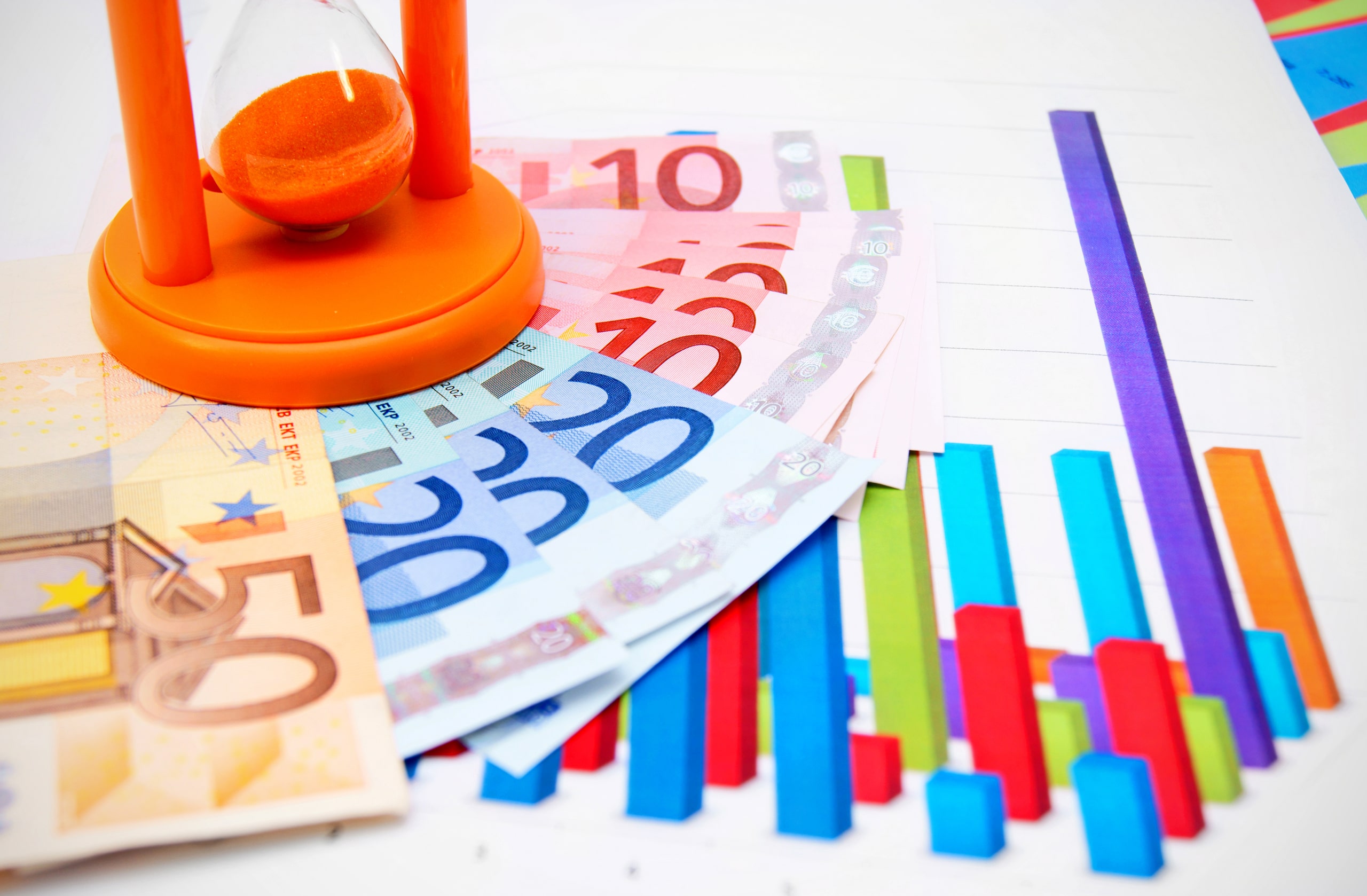 Rastreo De Operaciones Monetarias Con Gráficos Y Billetes De Euro Reloj de arena naranja sobre varios billetes de euro colocados sobre una hoja con gráficos de barras de colores que representan el análisis y rastreo de operaciones monetarias en finanzas y contabilidad.